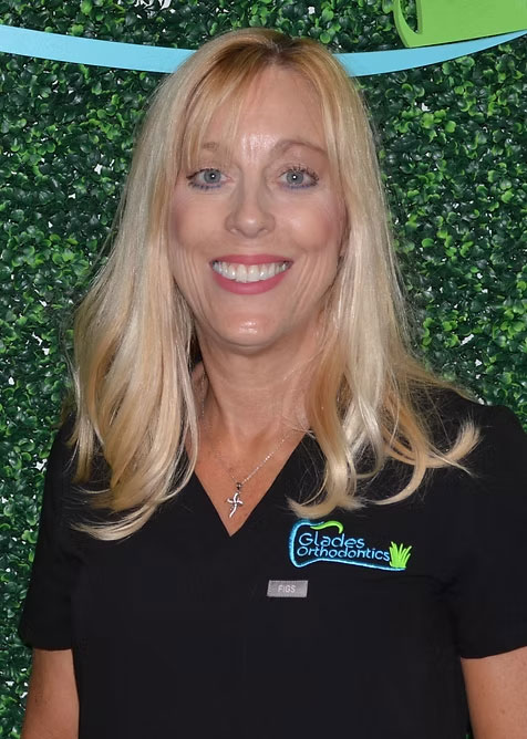 The image features a woman with blonde hair, wearing a black polo shirt with white lettering and a logo on the left chest area, standing against a green backdrop with a stylized border. She has a friendly expression and appears to be posing for the photograph.