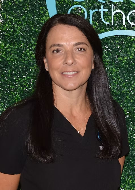 A woman with long dark hair wearing a white polo shirt stands against a green backdrop with a logo visible.