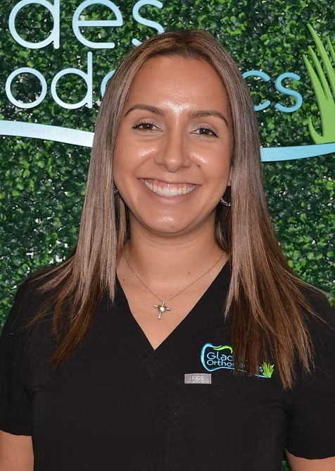 The image shows a woman wearing a dark-colored polo shirt with a logo on the left chest area, smiling at the camera, with a backdrop featuring a sign that reads  DES ODODOS.