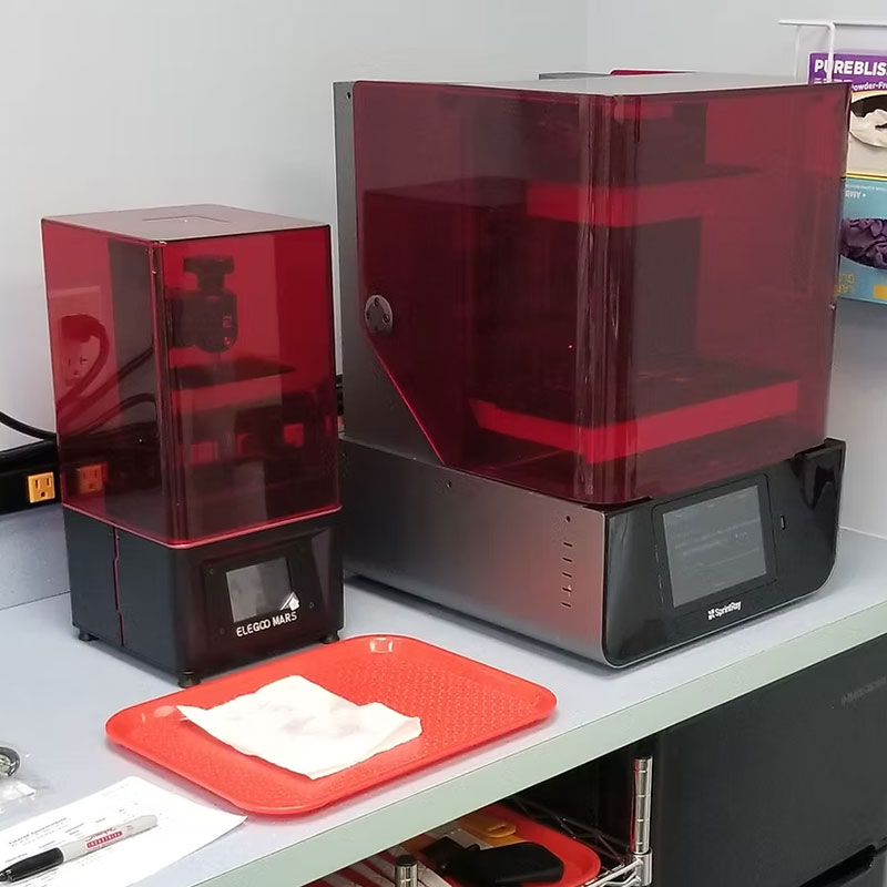 The image shows two 3D printers on a countertop with a red plastic cover partially removed from one printer, revealing its internal components.
